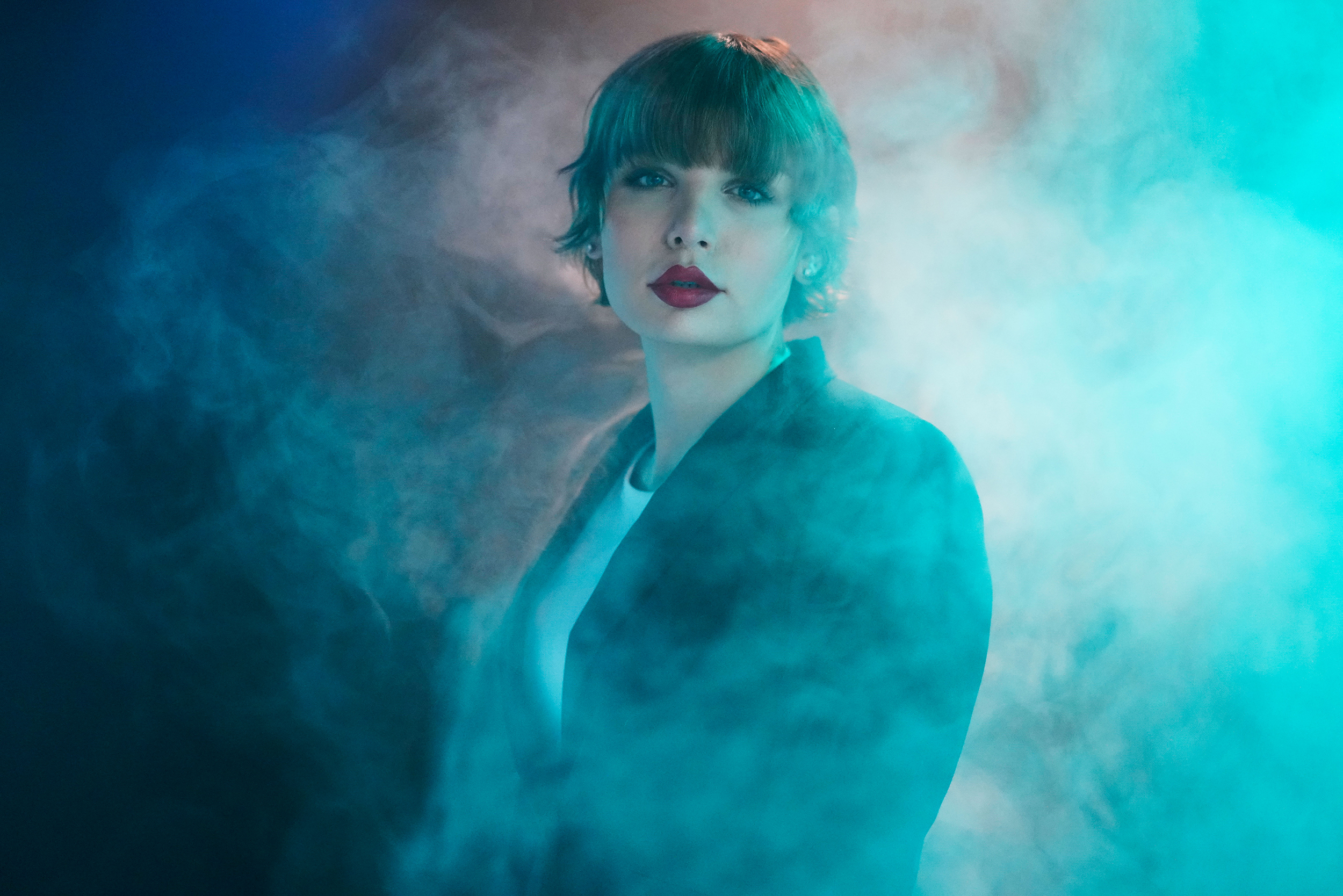 Aliée steht in schwarzem Blazer und weißem T-Shirt in grün-blauem Nebel und blickt in die Kamera. Die Lippen sind rot geschminkt.
