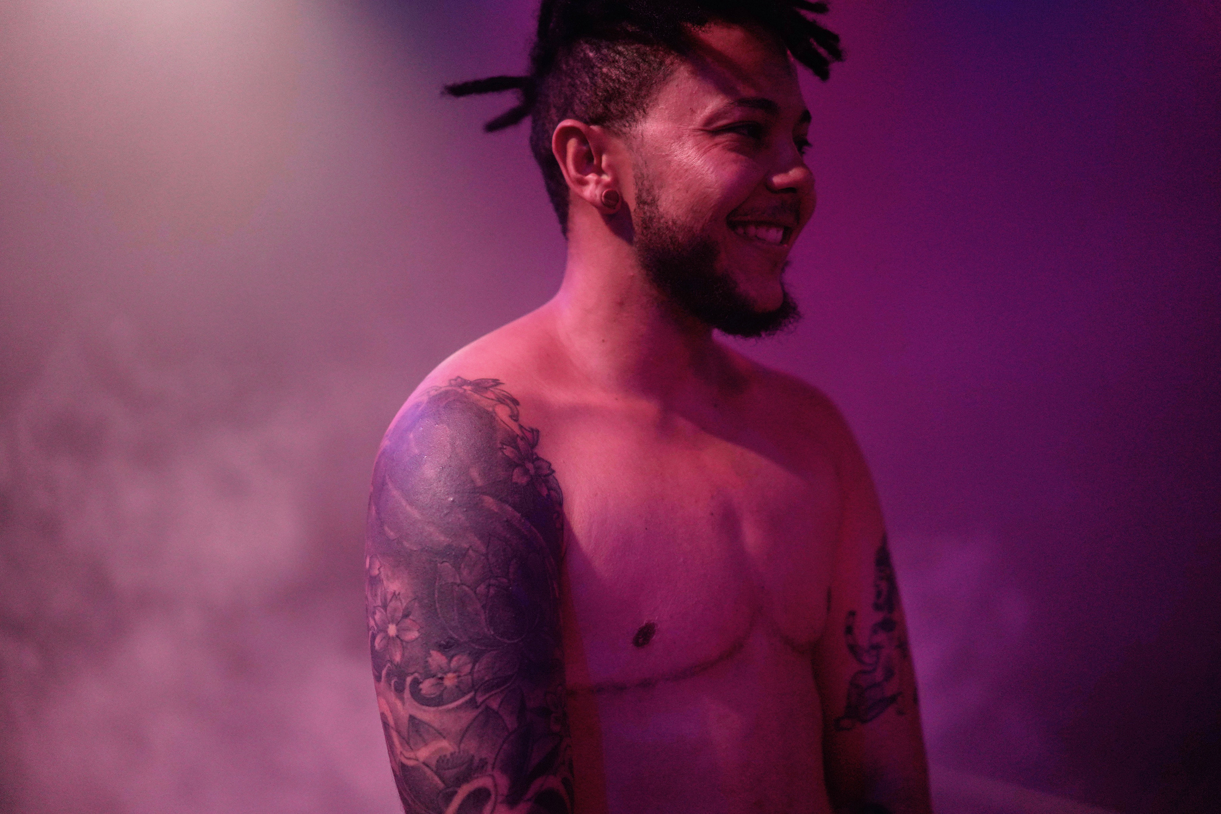 Bruno Santana steht oberkörperfrei in einem lila nebligen Raum und lacht. Man sieht Tattoos an den Armen und Narben auf der Brust.