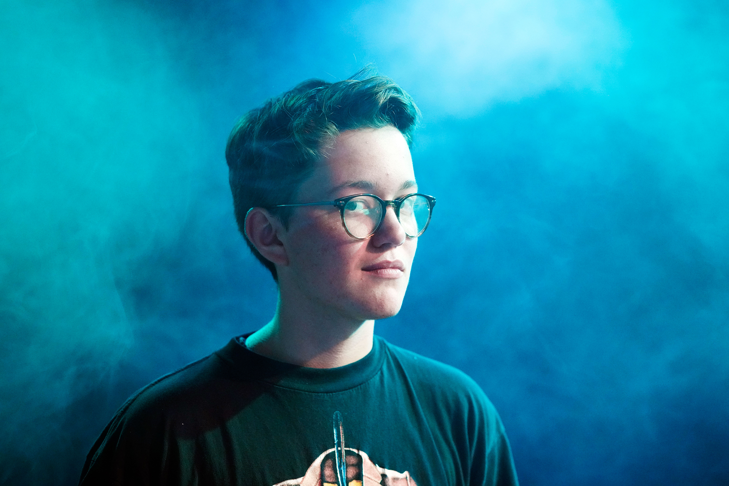 Jonathan mit kurzen braunen Haaren und Brille in einem blau nebligen Raum blickt in die Kamera.