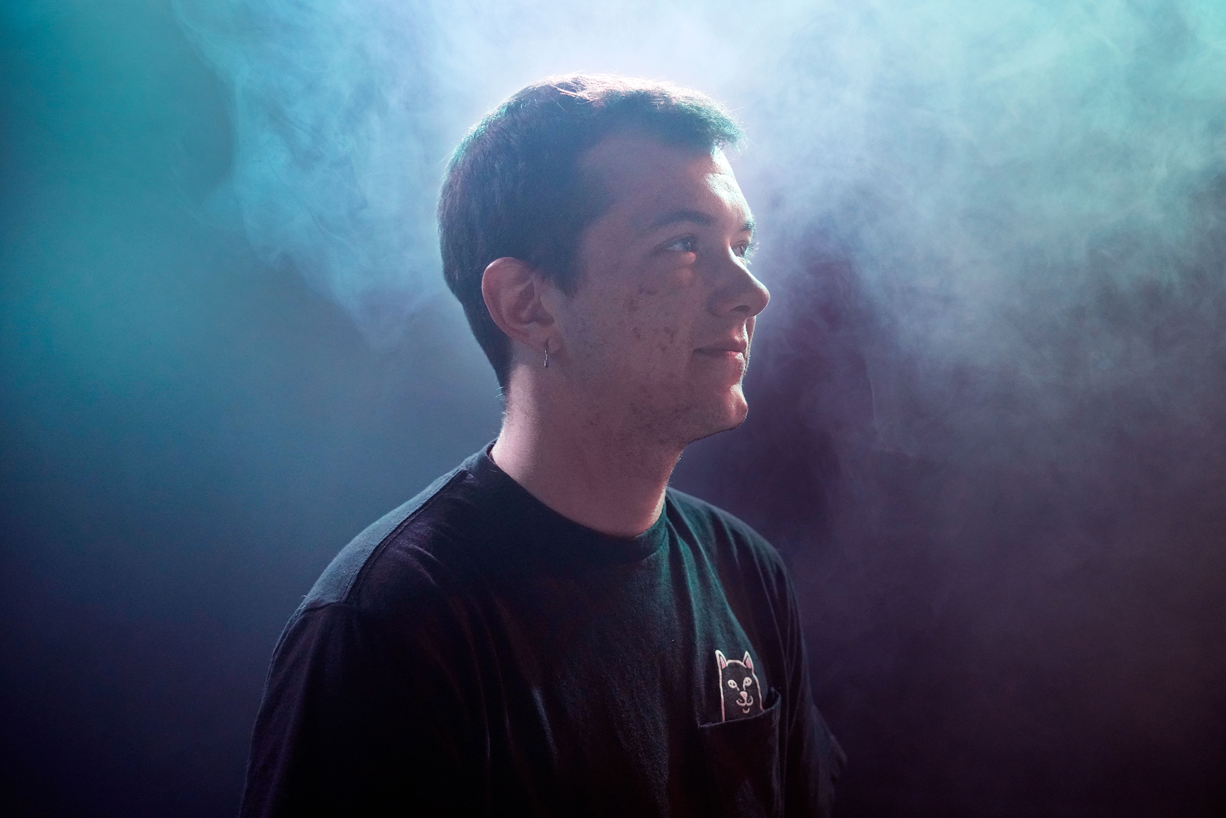 Lenny steht mit kurzen, braunen Haaren und schwarzem T-Shirt im Profil zur Kamera in einem dunklen Raum mit blauem Nebel.