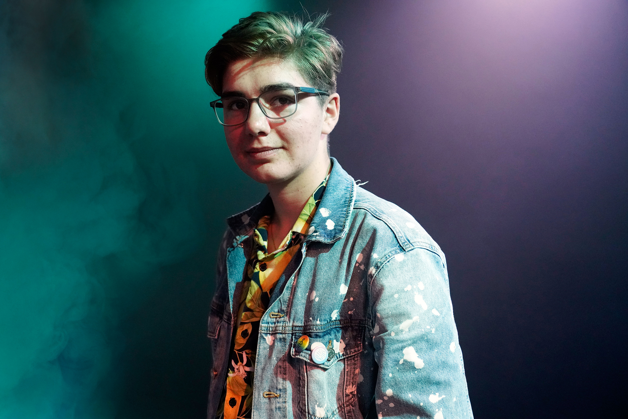 Luca steht mit kurzen, braunen Haaren und Jeansjacke seitlich zur Kamera in einem dunklen Raum mit lila-türkisem Nebel.