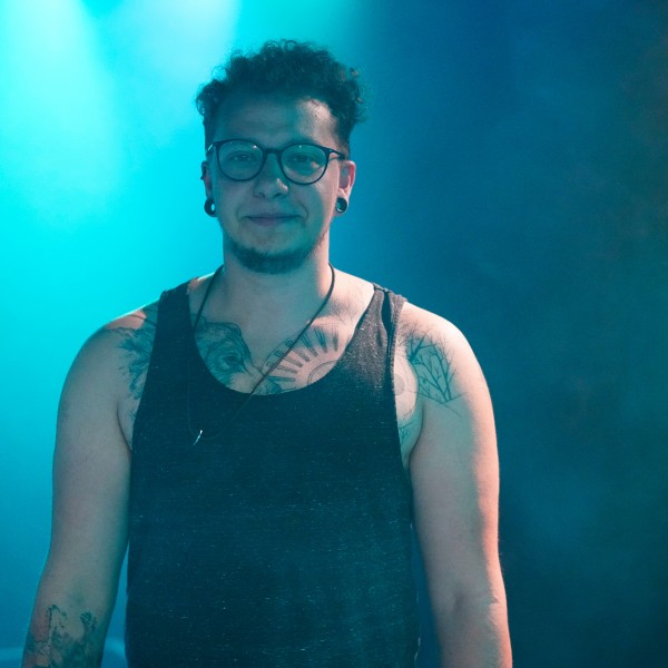 Philian stands in a turquoise purple foggy room with short curly hair wearing glasses and a black vest top. Tattoos are shown on the chest and arms.