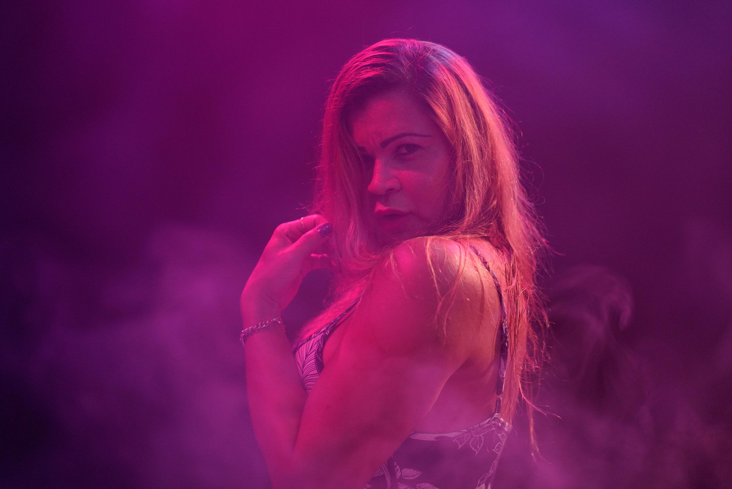 Ranella steht im Profil, ein Kleid mit schmalen Schulterriemen tragend, in einem dunkelvioletten, nebligen Raum mit langen blonden Haaren und schaut über ihre linke Schulter in die Kamera.