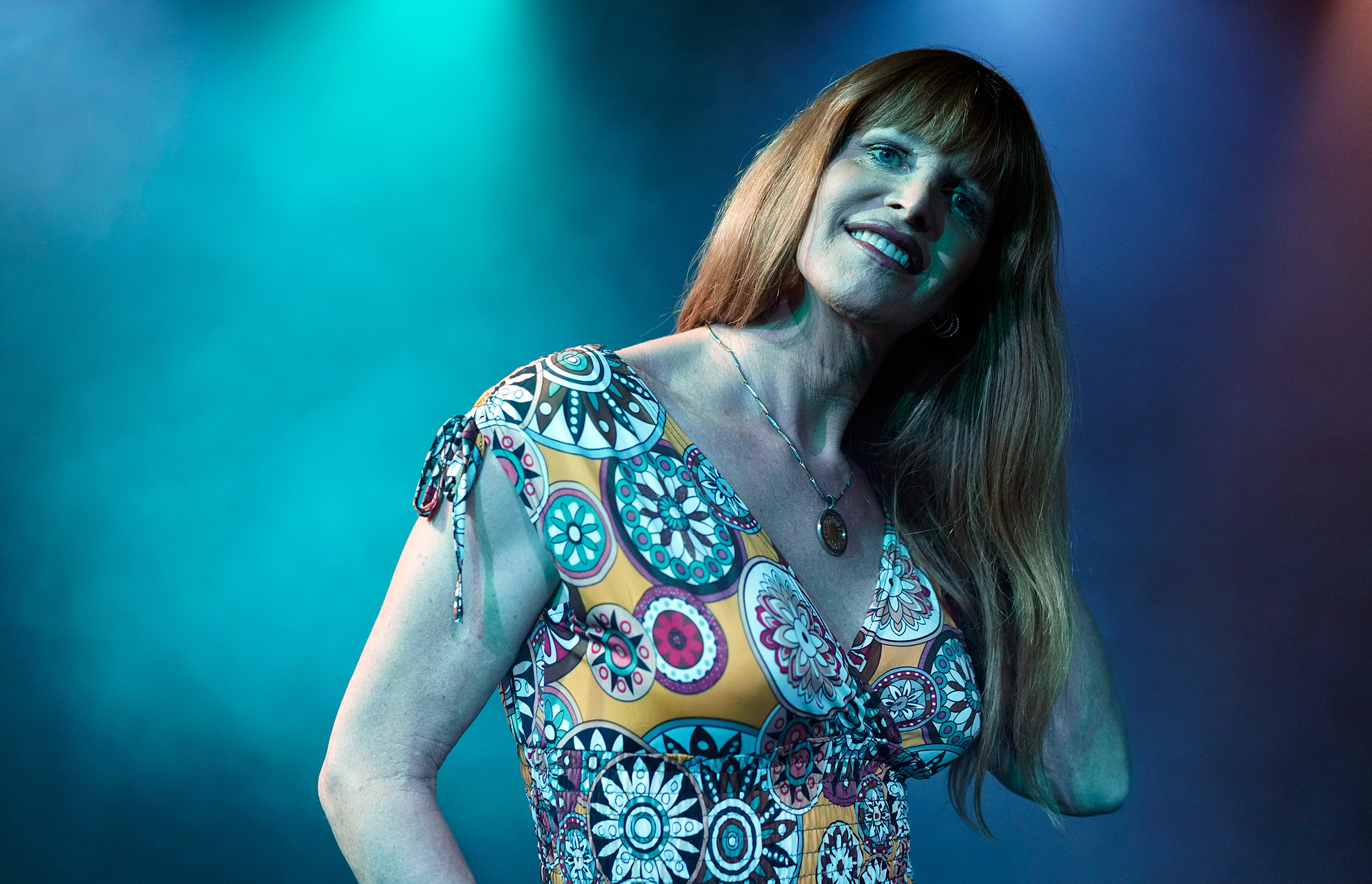 Claudia steht mit rötlichen, brustlangen Haaren in einem Blumenkleid in einem blau nebligen Raum.