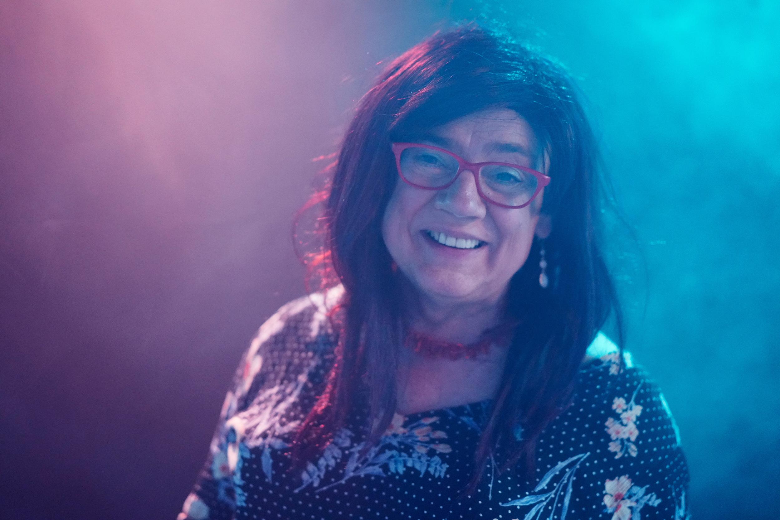 Sonja Elaine steht lächelnd mit rotbraunem Haar in einem türkisvioletten Nebelraum, trägt eine Brille, eine rote Halskette und ein blumiges Kleid.