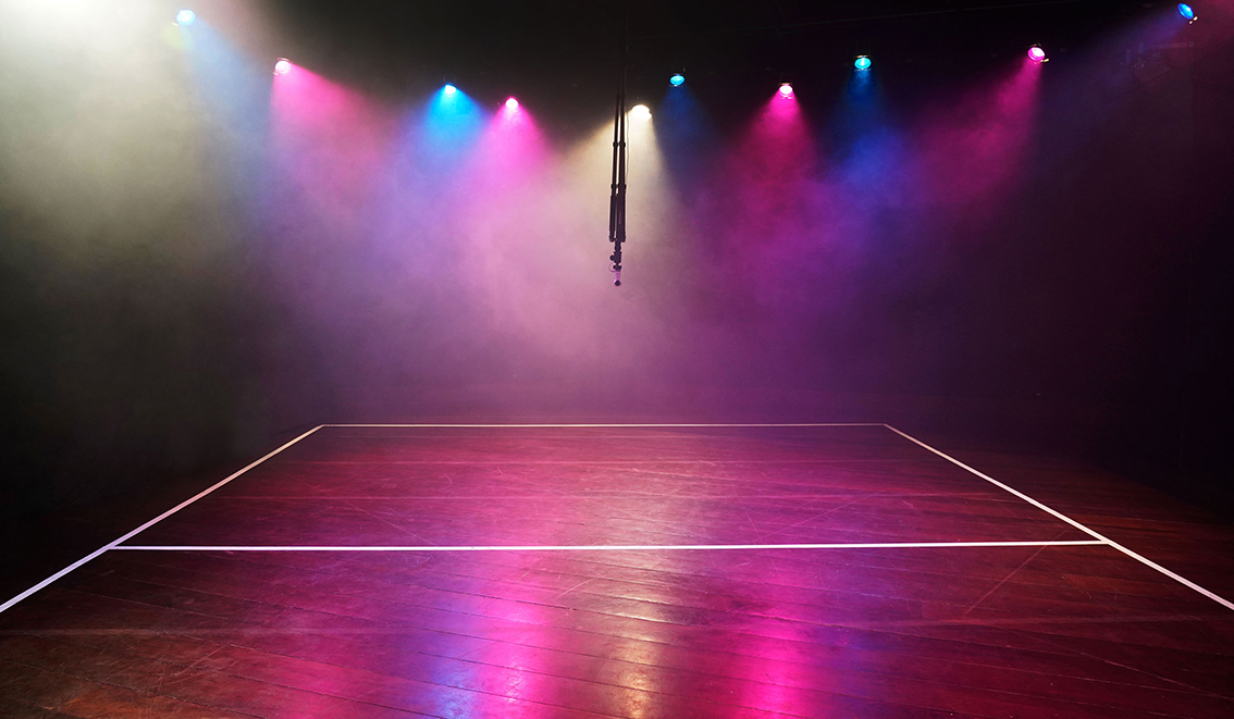 Ein dunkler, menschenleerer Raum mit einer Mischung aus rosa-blauen und weißen Theaterlichtern, die Nebelwolken erhellen. Die Lichter spiegeln sich auf dem Holzboden. Die Bodenlinien für eine Mannschaftssportart sind auf dem Boden mit weißem Klebeband markiert. In der Mitte des Raumes hängt eine 360-Grad-Kamera.