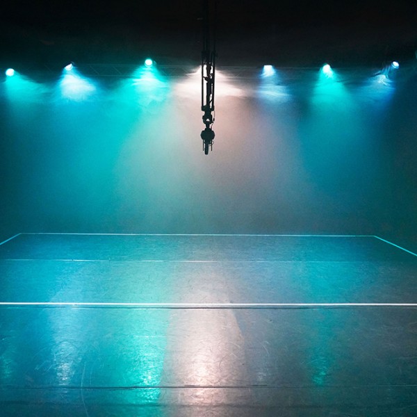 A dark room empty of people with a mixture of turquoise, blue and white theatrical lights that are illuminating clouds of fog. The lights are reflected on the floor. The boundaries for a team sport are marked out on the floor with white tape. Hung in the middle of the room is a 360 degree camera.