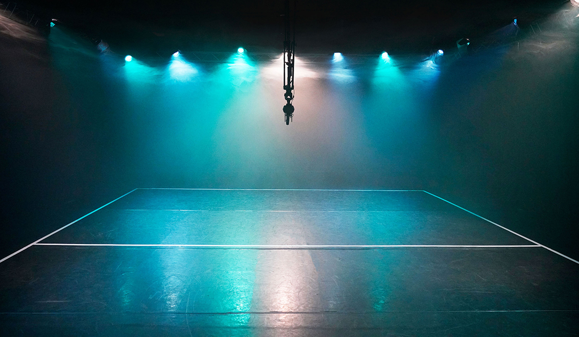 Ein dunkler, menschenleerer Raum mit einer Mischung aus türkisfarbenen, blauen und weißen Theaterlichtern, die Nebelwolken erhellen. Die Lichter werden auf dem Boden reflektiert. Die Grenzen für eine Mannschaftssportart sind auf dem Boden mit weißem Klebeband markiert. In der Mitte des Raumes hängt eine 360-Grad-Kamera.