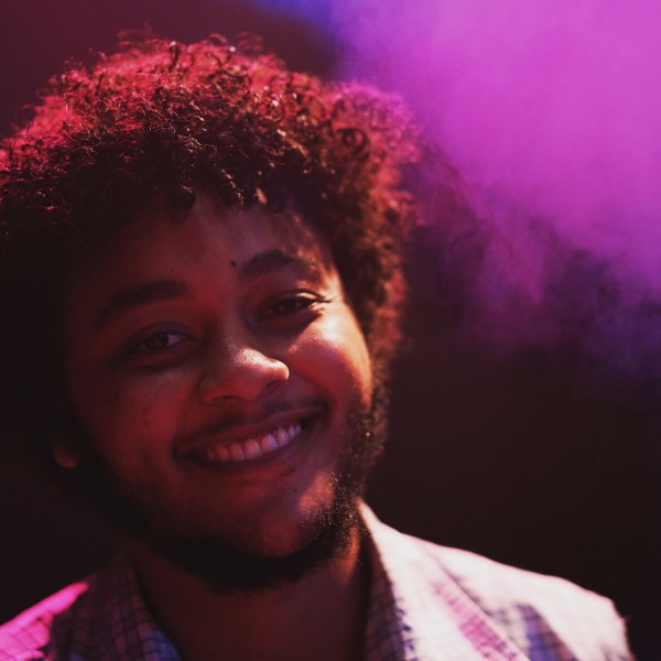 Theo Brandon with short dark brown coiled hair and beard stands smiling in close up in a dark purple foggy room.