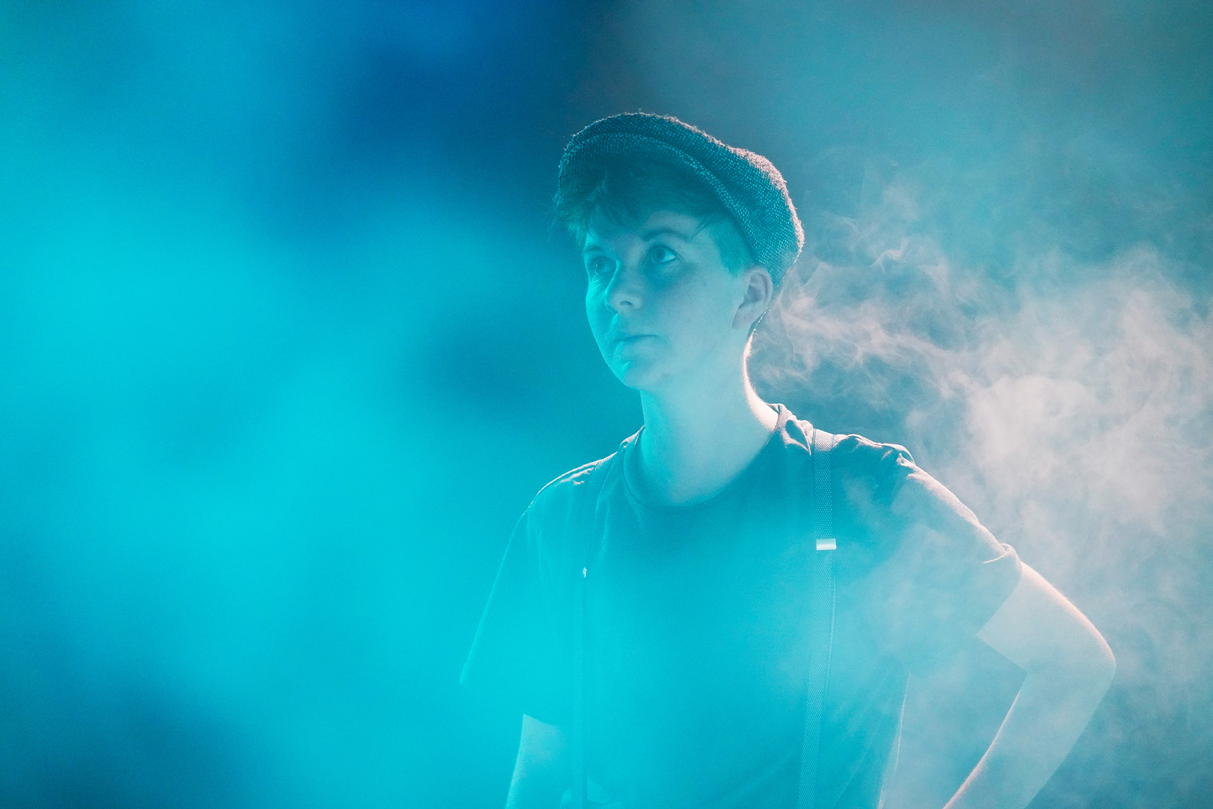 Tjark steht in einem blauvioletten Nebel, trägt eine flache Mütze, T-Shirt und Hosenträger und blickt nach oben und rechts.
