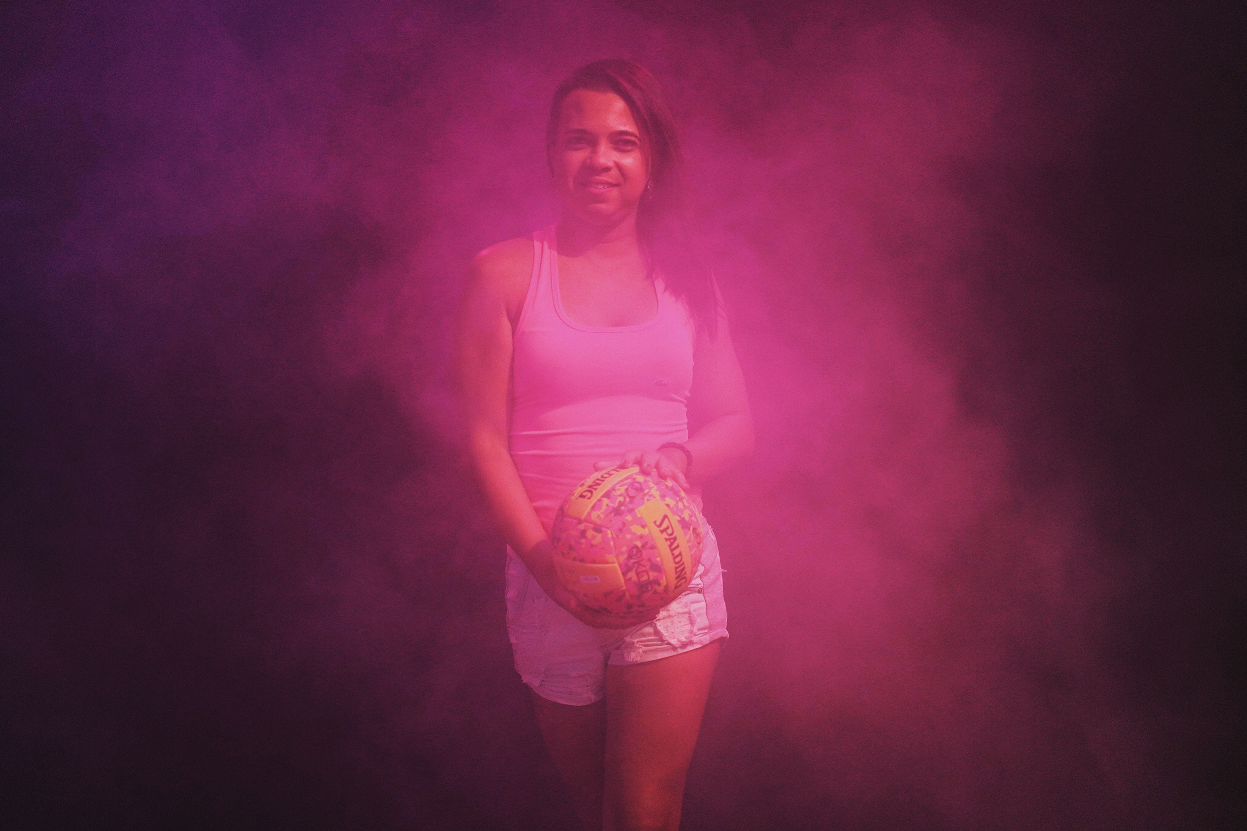 Viviane in Shorts und ärmellosem Oberteil steht lächelnd, teilweise verdeckt, in einem dunkelvioletten, nebligen Raum und hält einen Ball auf Hüfthöhe in den Händen.